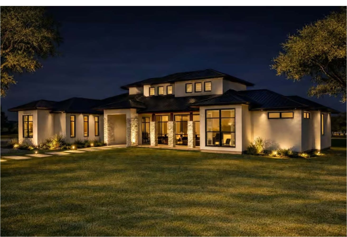 The Monaco — single-story estate at night with light stucco walls, stone entry columns, a dark low-pitch roofline and mature oak trees